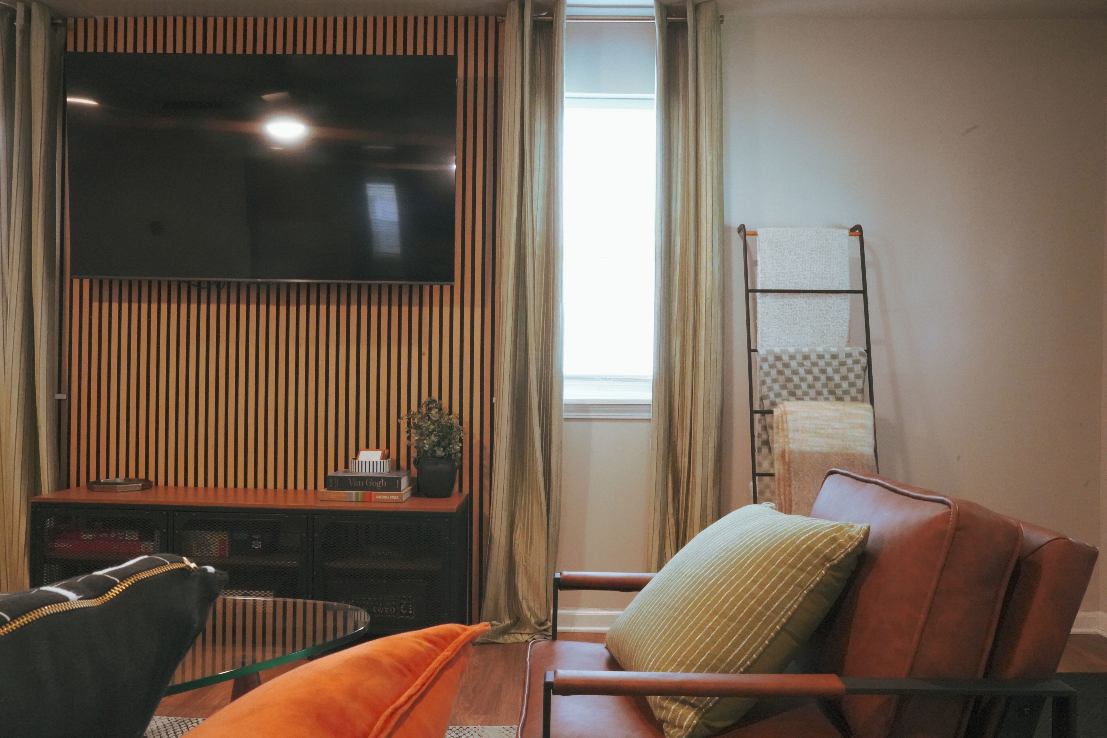 Un salón con una televisión de pantalla plana montada en una pared de madera, un sofá con cojines, una mesa de centro de vidrio y una ventana con cortinas.