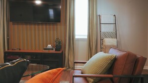 A living room with a flat-screen TV mounted on a wooden wall, a sofa with cushions, a glass coffee table, and a window with curtains. - Spacious, Sleeps 16+, Game Room Near Downtown Indianapolis (Indianapolis)