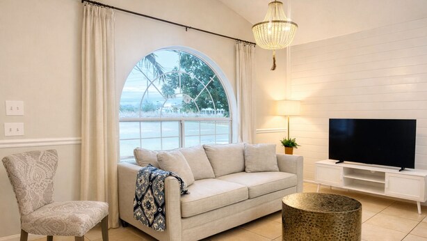 Ein Wohnzimmer mit einer Couch, einem Sessel, einem Fernseher, einer Lampe und einem großen Fenster mit Vorhängen.