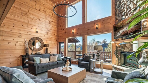 A cozy living room with wooden walls, a fireplace, and a large window overlooking a scenic view. - VIEWS! Sleeps 22 | Pool | Theater | Game Rm | (Sevierville)