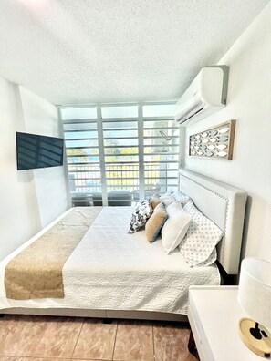 A bedroom with a large bed, a TV mounted on the wall, and a window with blinds. - Beachfront 2-Bedroom Apartment with Resort-Style Amenities in Luquillo (Luquillo)