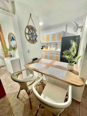 A dining area with a wooden table, two wicker chairs, a potted plant, and a kitchen in the background. - Beachfront 2-Bedroom Apartment with Resort-Style Amenities in Luquillo (Luquillo)