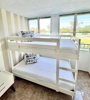 A bunk bed with white bedding and patterned pillows, a small white desk, and a window with a view of palm trees and a sports field. - Beachfront 2-Bedroom Apartment with Resort-Style Amenities in Luquillo (Luquillo)