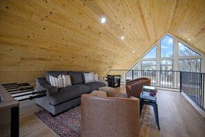 A loft-style room with a wooden ceiling, a gray sofa, a coffee table, and a view through large windows. - Modern Black Woods Creek Cabin (McCurtain County)