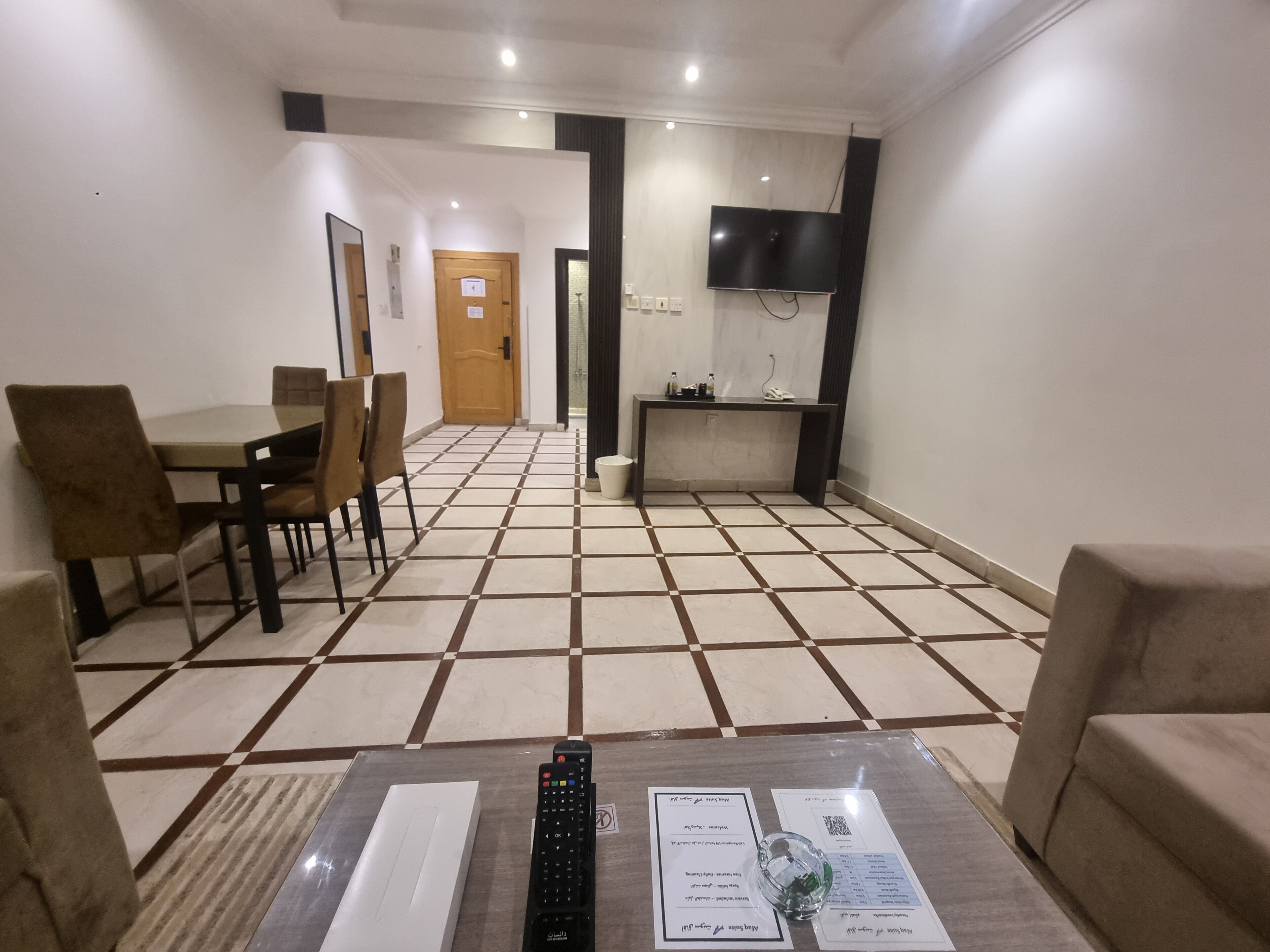 Una habitación de hotel con piso de baldosas, una mesa de comedor con sillas, un televisor montado en la pared y un escritorio pequeño con un teléfono.