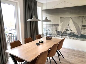 A dining area with a wooden table and brown chairs, a large window with curtains, and a mural of a sailing ship on the wall. - Beautiful Lodge Located on the Church Square (Zoutelande)