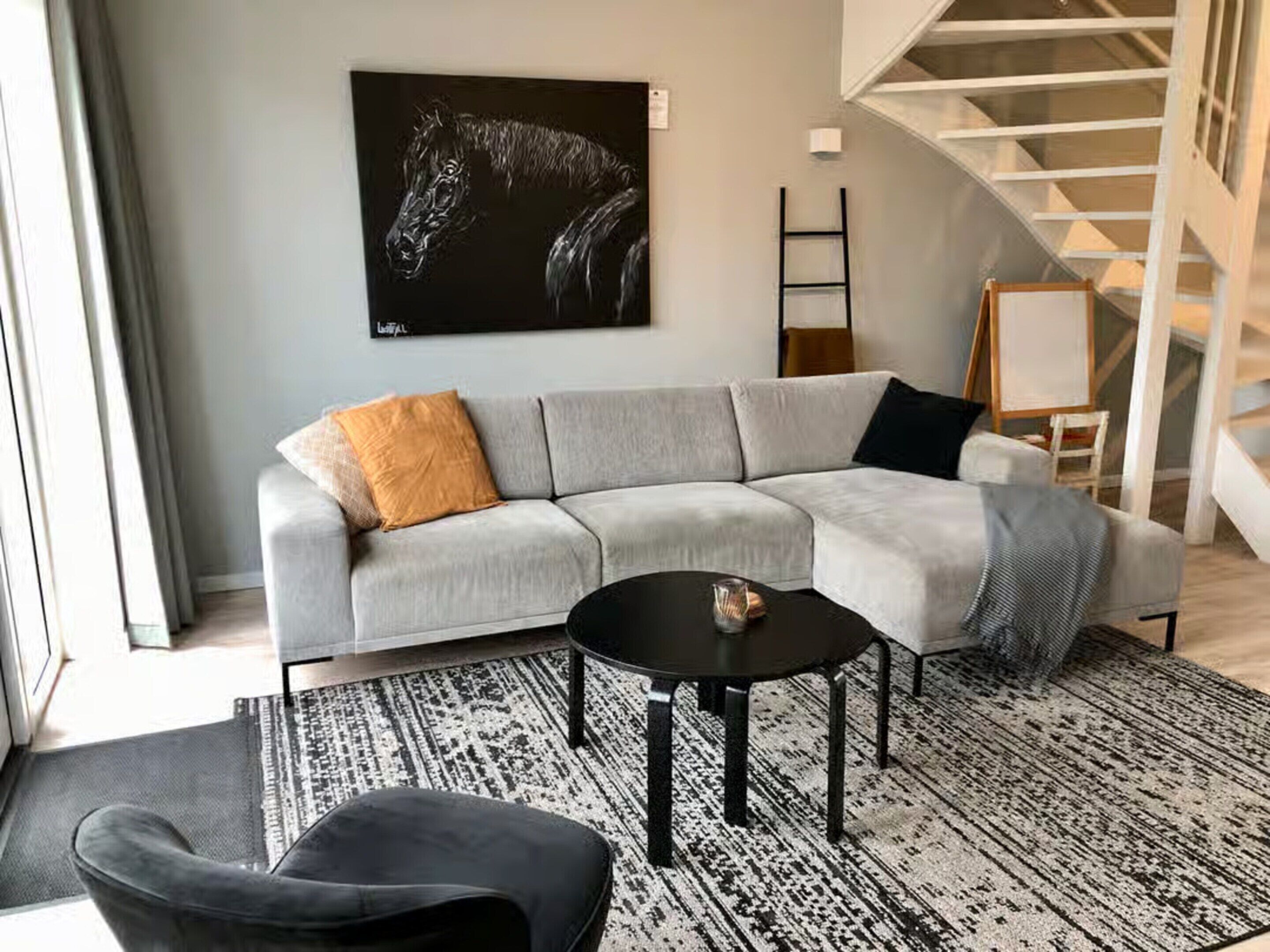 A modern living room with a grey sectional sofa, a black coffee table, and a black armchair. A black and white patterned rug covers the floor. A large black painting of a horse is on the wall.