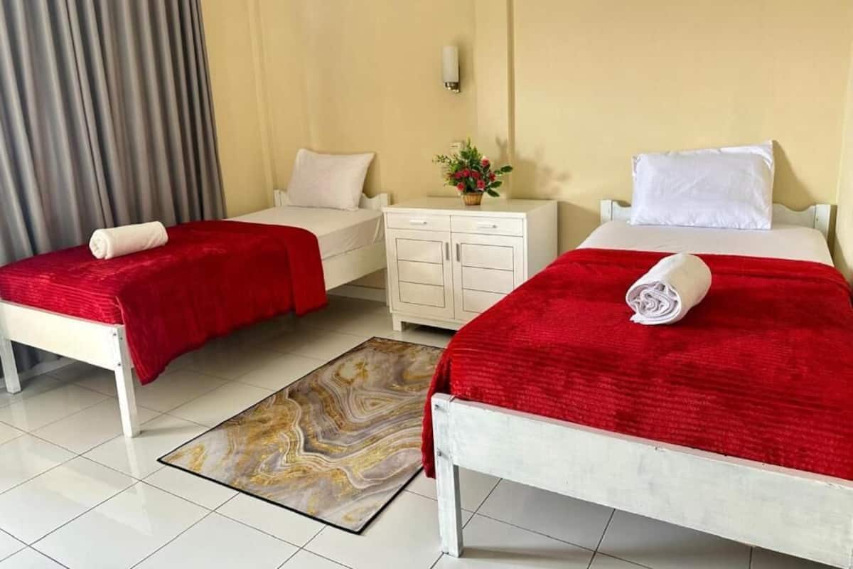 Habitación de hotel con dos camas, cada una con una colcha y almohadas rojas, una mesita de noche blanca con un jarrón de flores y una alfombra estampada con diseño de mármol.