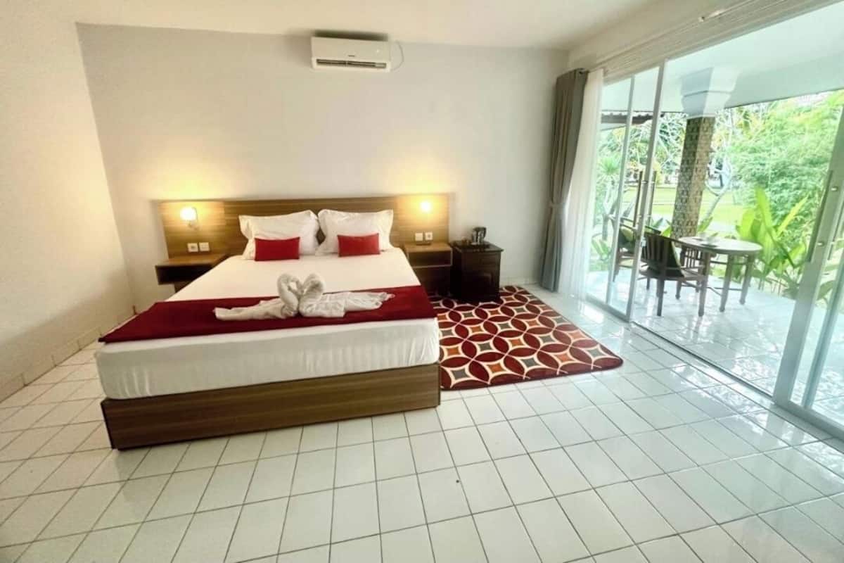 Habitación de hotel con una cama grande, una mesita de noche, una ventana con vistas a la vegetación y una puerta corrediza de vidrio a un espacio exterior.