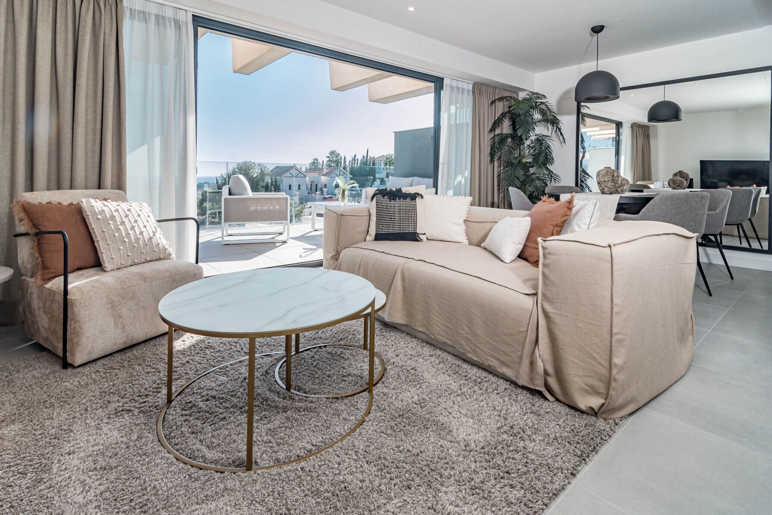 A modern living room with a beige sofa, a round coffee table, and a large window offering a view of the city.