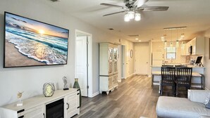 A spacious living room with a large ocean view painting, a ceiling fan, and a modern kitchen with pendant lights. - Forest Park – 2BR Pool, Clubhouse & Beaches (Englewood)