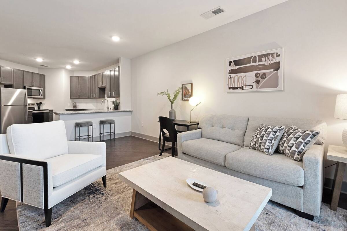 A modern living room with a grey sofa, a white armchair, and a coffee table. There is a kitchen in the background with grey cabinets and stainless steel appliances.