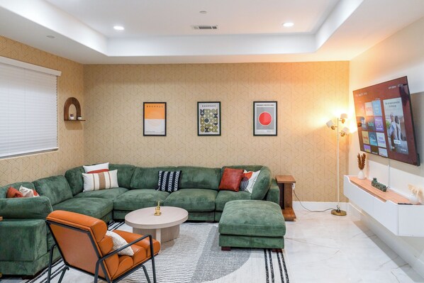 Een moderne woonkamer met een groene hoekbank, een ronde salontafel en een flatscreen televisie aan de muur.