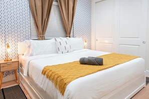 Een net opgemaakt bed met een witte dekbedovertrek en een mosterdgele plaid, een bedlampje en een nachtkastje met een gestreept lantaarn.