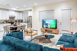 Een moderne woonkamer met een grote flatscreen televisie, een bank, een salontafel en een eethoek met stoelen.