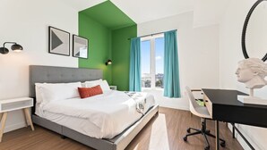 Ein modernes Schlafzimmer mit grauem Bett, weißer Bettwäsche, einer grünen Akzentwand, einem Schreibtisch mit Stuhl und einer Skulptur auf dem Schreibtisch.