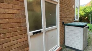 Une maison en briques avec une porte et des fenêtres blanches, un petit abri de jardin et un toit vert.