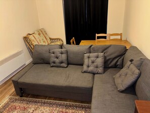 En grå hörnsoffa med två kvadratiska kuddar, en mönstrad matta, ett träbord och en stol.