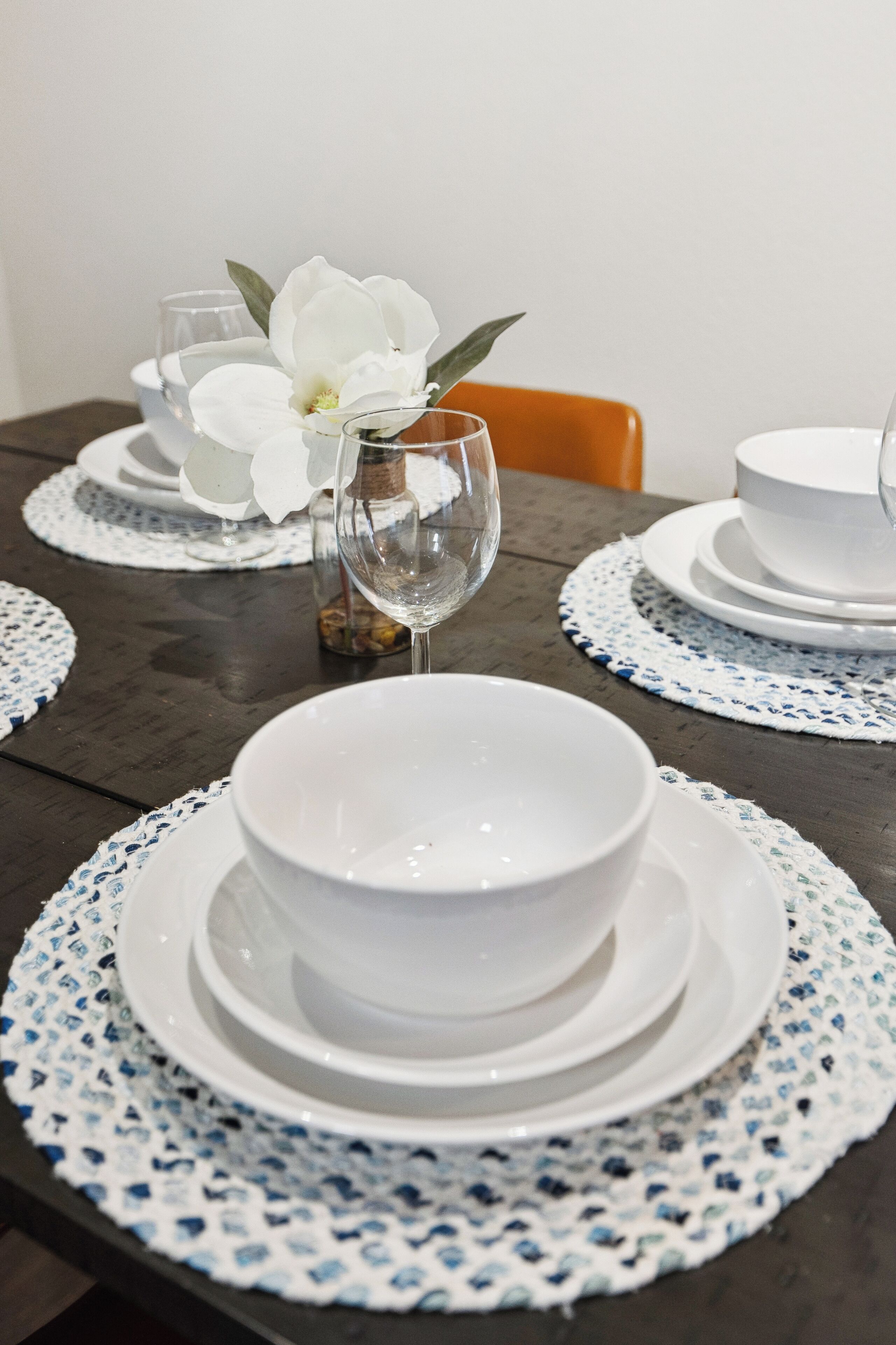 A dining table set with white plates, bowls, and a wine glass, accompanied by a white flower and a small vase.