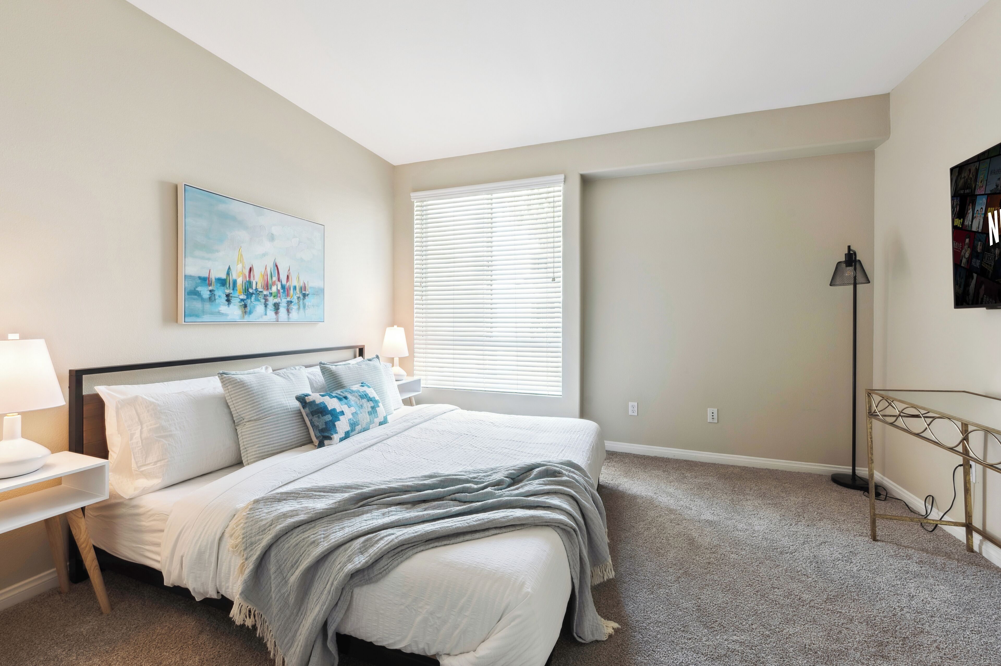 A bedroom with a bed, nightstands, a lamp, a painting, a window with blinds, and a floor lamp.