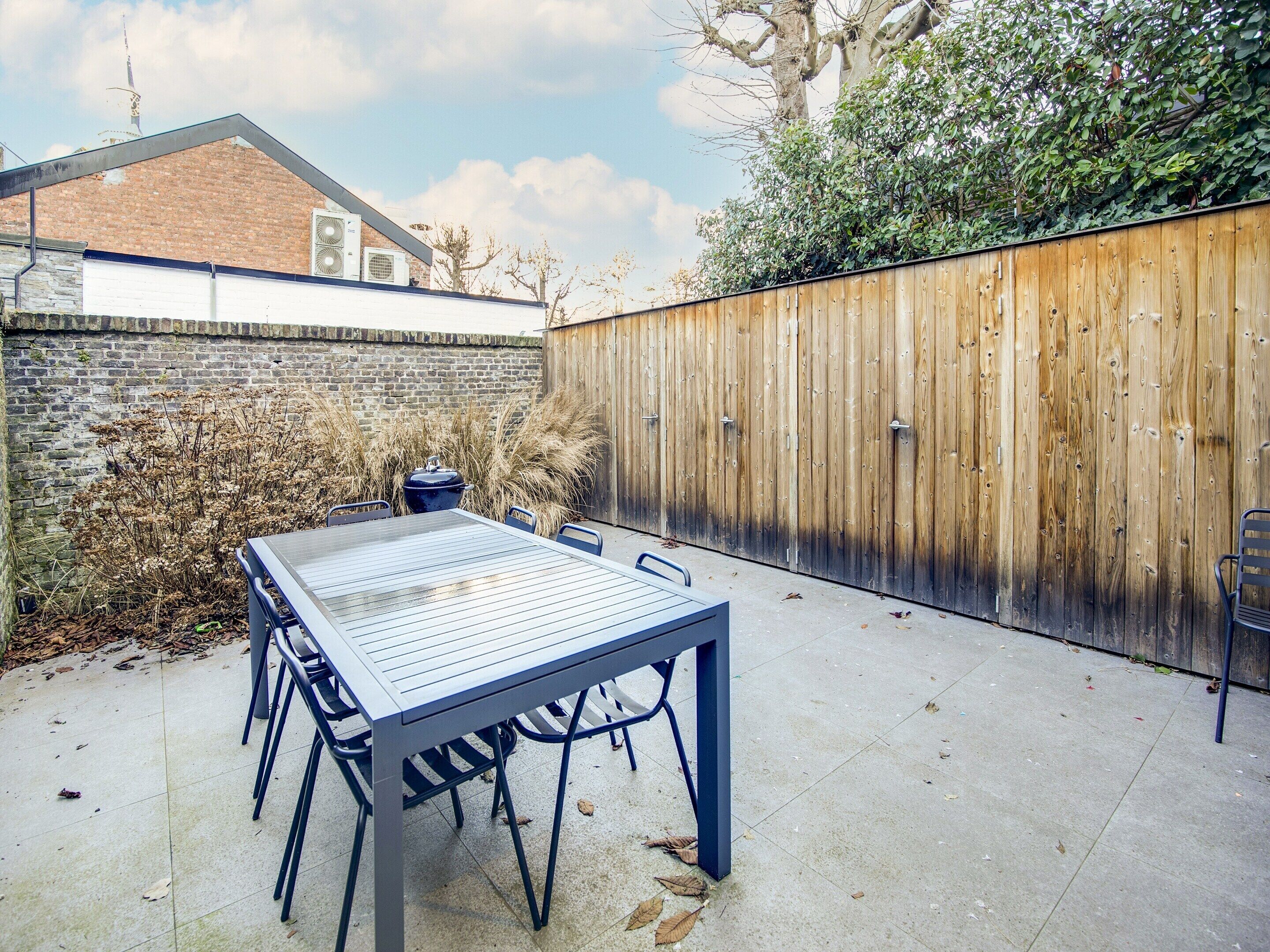 A patio with a table and chairs, a barbecue grill, a wooden fence, and a brick wall.