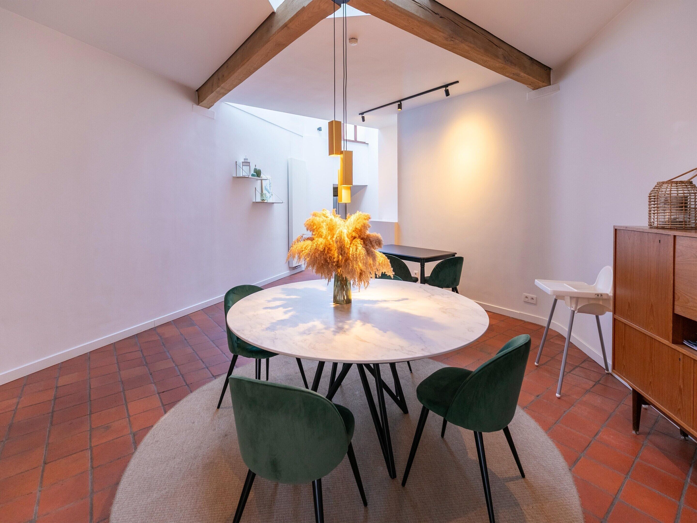 Un coin repas avec une table ronde, des chaises vertes et un vase contenant des fleurs séchées.
