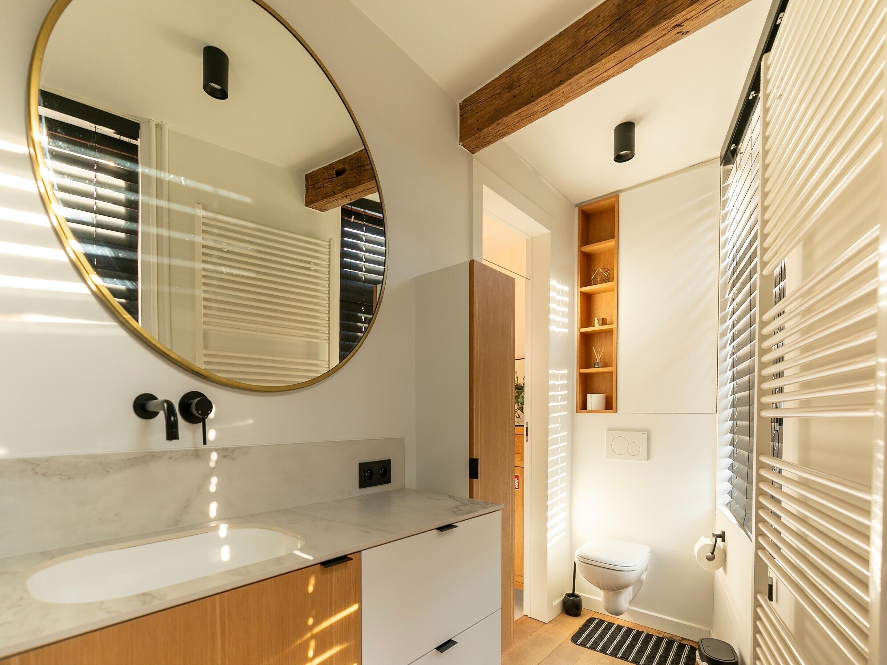 A modern bathroom with a large mirror, wooden beams, a built-in shelf, and a toilet with a radiator.
