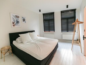 A bedroom with a black bed, white bedding, two windows with blinds, a wooden stool, and abstract wall art. - Holiday Home in the Heart of Hasselt (Hasselt)
