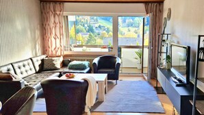 A living room with a sofa, armchair, and a table with a bowl of fruit. There is a large window with a view of a green landscape. - House Sunshine on the ski Slope (Herrischried)