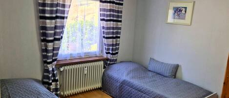 Un alojamiento pequeño y de una sola habitación con dos camas individuales, un radiador y una ventana con cortinas.