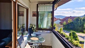 A balcony with a table and chairs, a window with lace curtains, and a view of houses and greenery. - House Sunshine on the ski Slope (Herrischried)