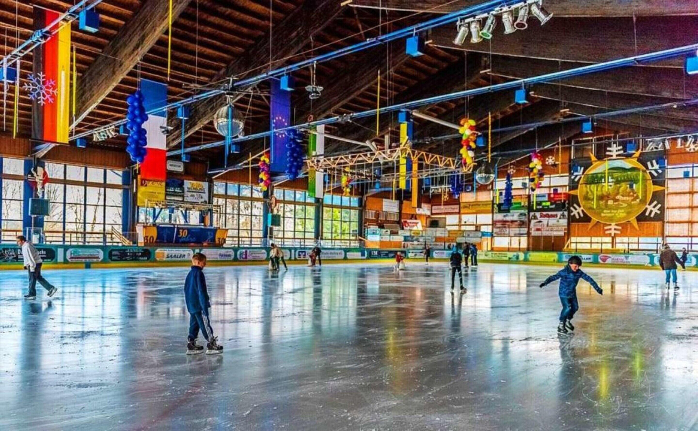 An indoor ice skating rink with multiple skaters, wooden beams, and colorful banners.