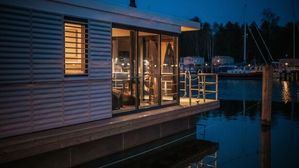 A waterfront house with a glass door leading to a deck, a fire pit, and a dock extending into the water. - Floating Houseboat in Peenemunde (Peenemünde)