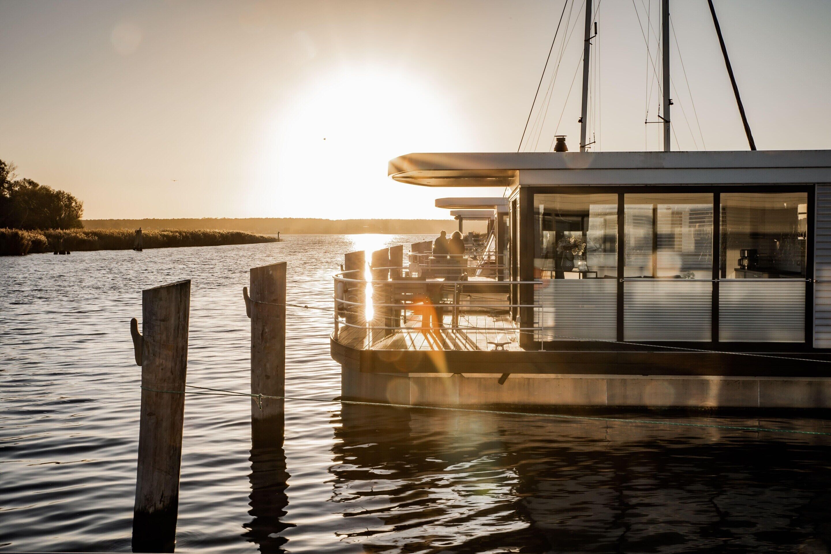 A boat with a covered deck and large windows, docked at a pier with calm water and a clear sky.