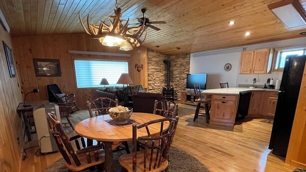 A cozy cabin interior with a wooden dining table surrounded by chairs, a stone wall, a stone fireplace, and a kitchen area with wooden cabinets and a black refrigerator. - Mountain View Barndominium at Black Bear Ranch (Nordic)