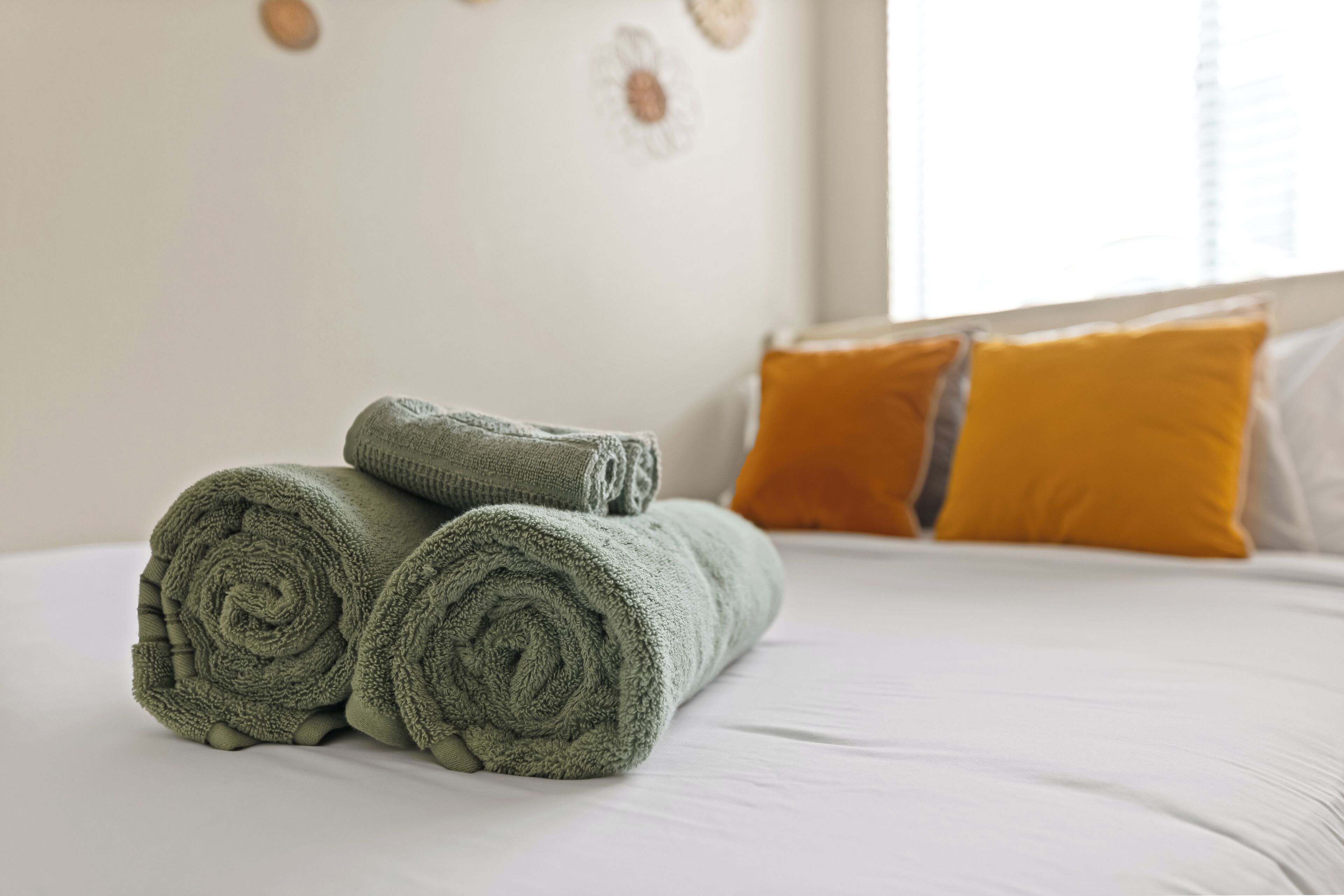 A neatly made bed with rolled green towels and a single orange pillow.