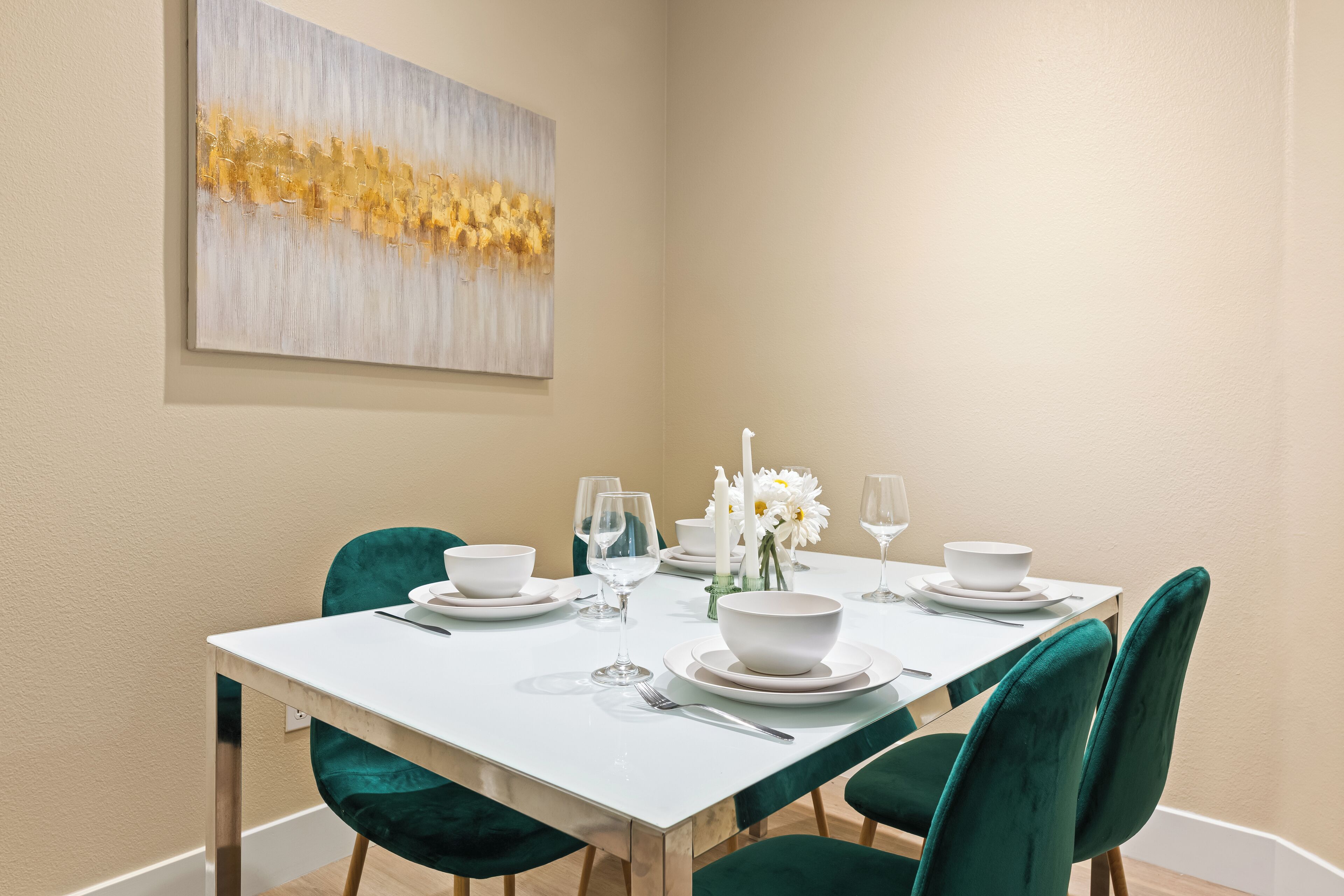 Un coin repas avec une table blanche dressée pour quatre personnes, des chaises en velours vert, des assiettes blanches, des bols et des verres à vin. Un vase blanc rempli de fleurs est posé sur la table, et un tableau orne le mur.