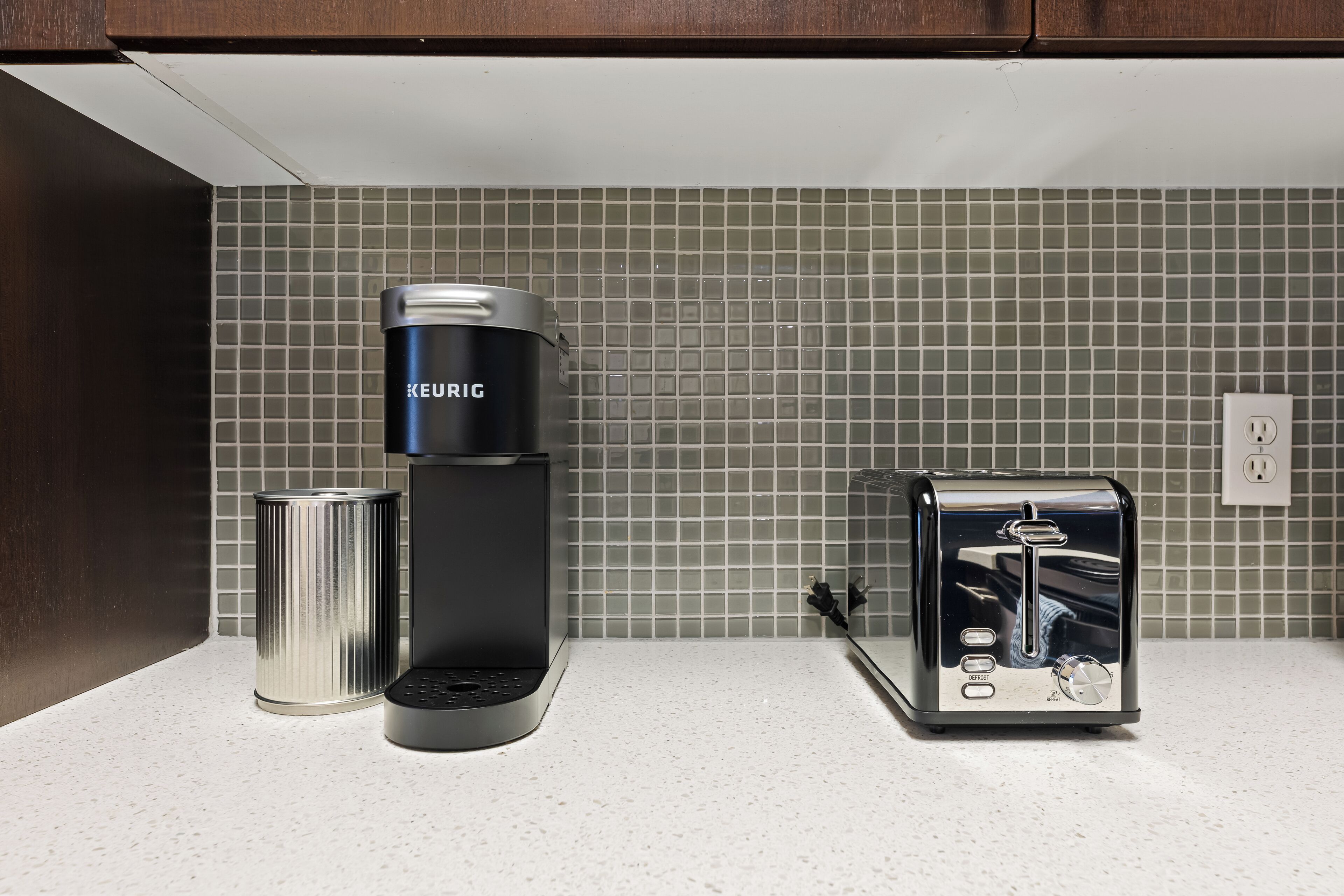 Une cuisine équipée d’une machine à café Keurig, d’un grille-pain et d’une boîte en acier inoxydable, le tout posé sur un plan de travail blanc.