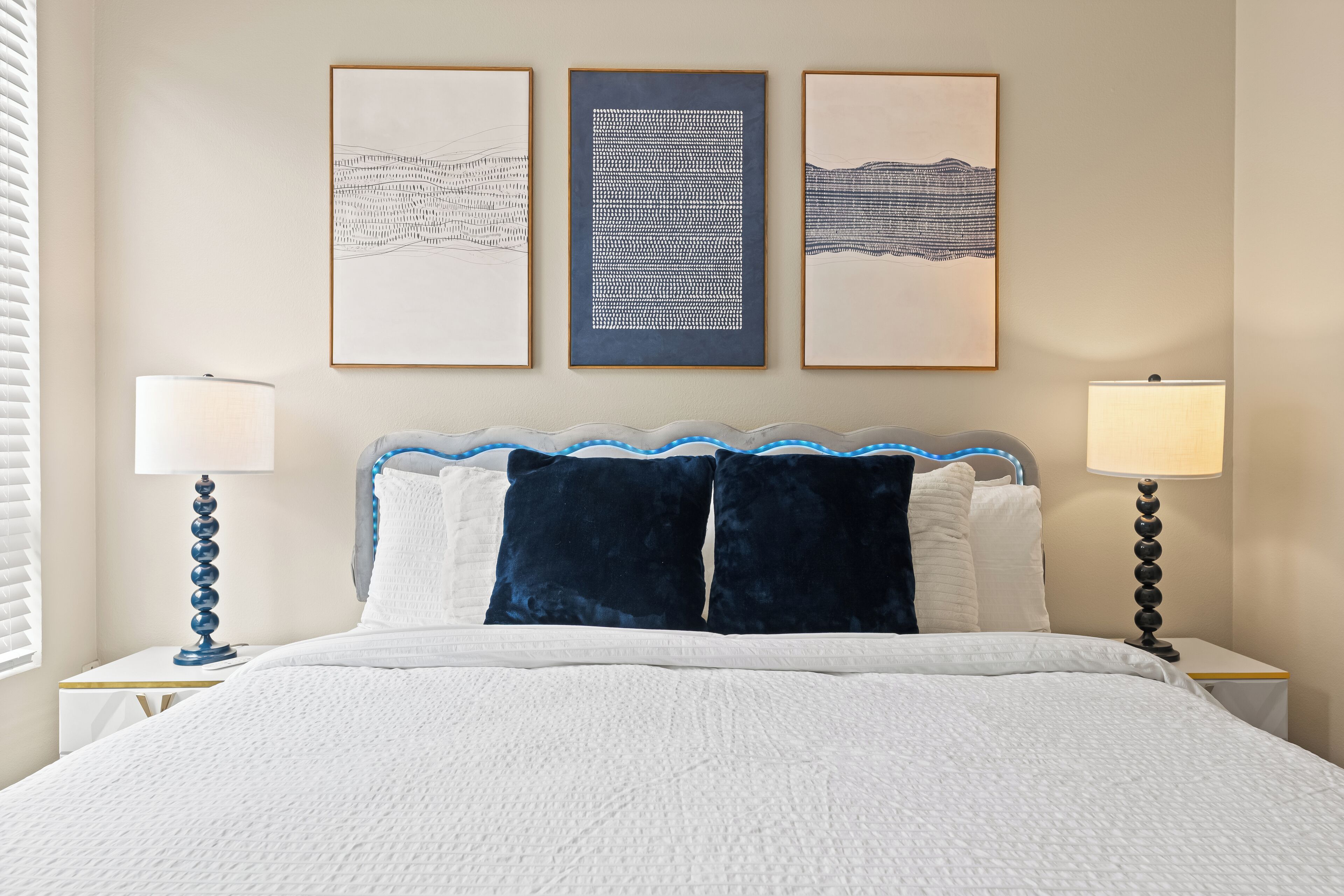 A neatly made bed with a white and blue headboard, two bedside lamps, and three framed artworks on the wall.