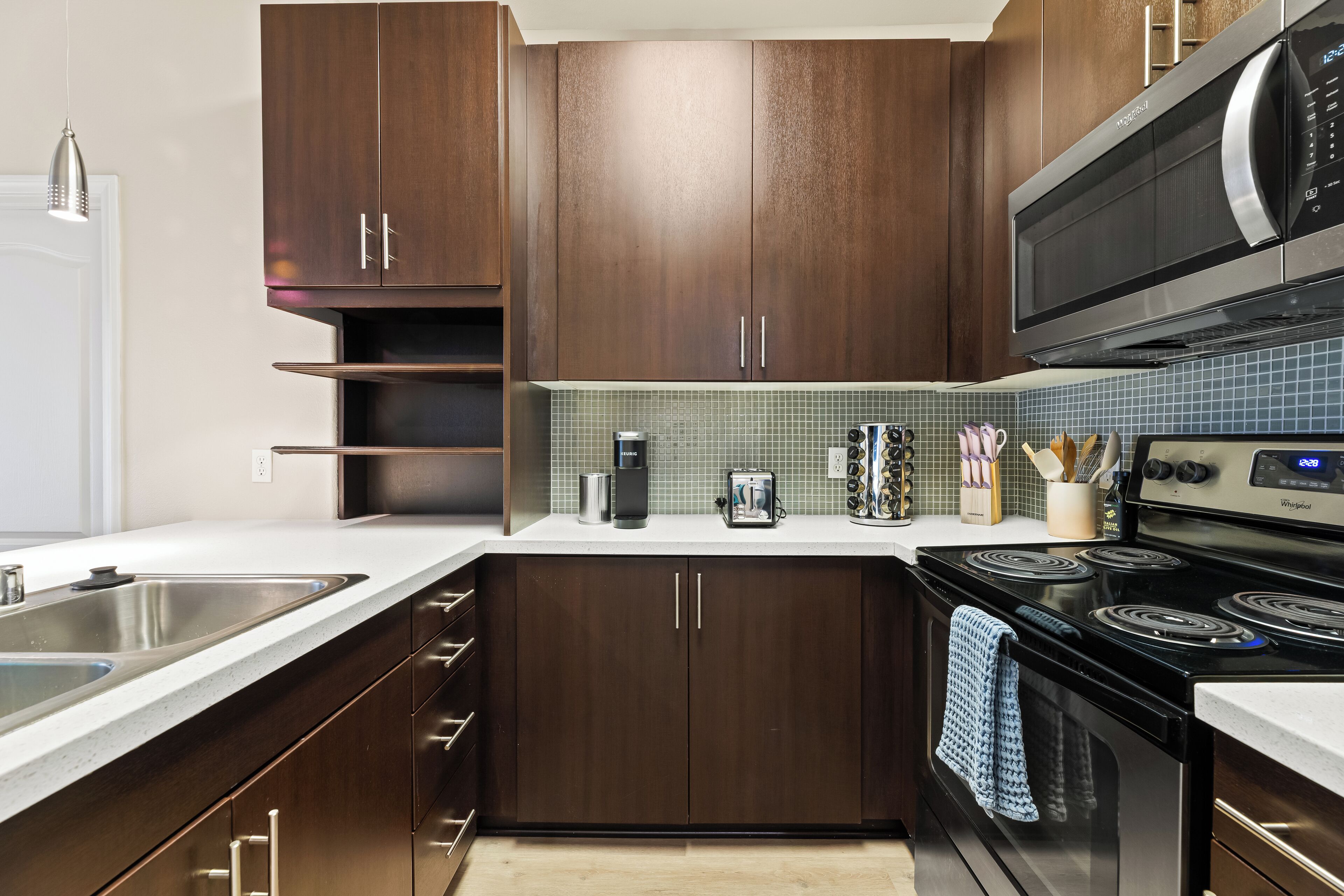 A modern kitchen with dark wooden cabinets, a stainless steel sink, a microwave, a stove, and a tiled backsplash.