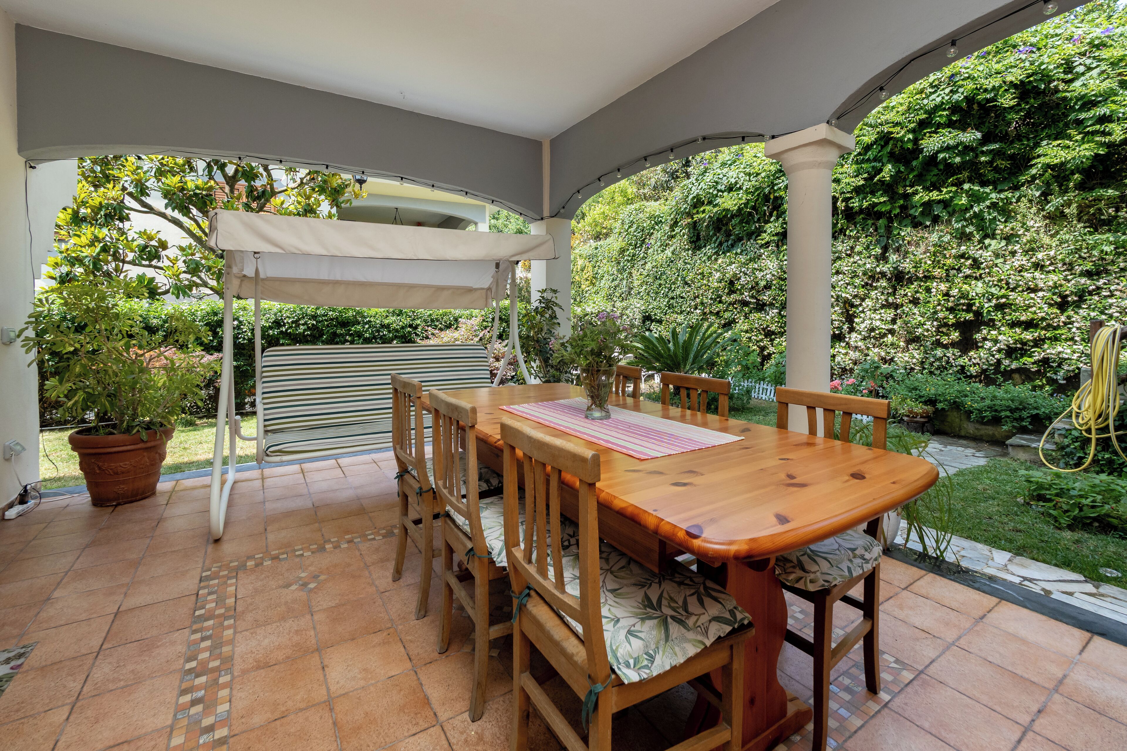 Una veranda riparata con tavolo e sedie in legno, circondata da piante in vaso e un giardino.