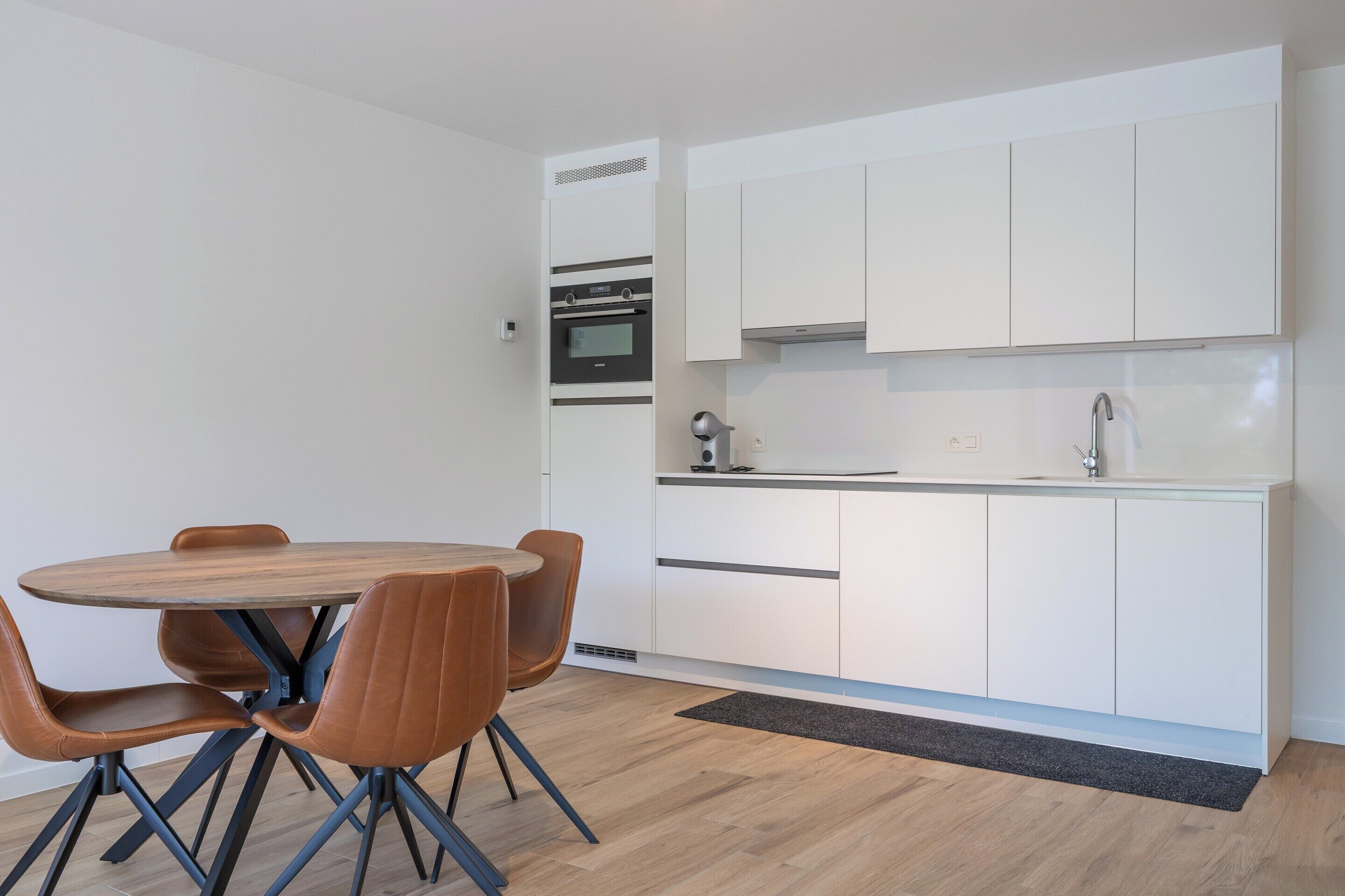 Una cocina moderna con armarios blancos, un horno integrado y una mesa redonda de madera con cuatro sillas.