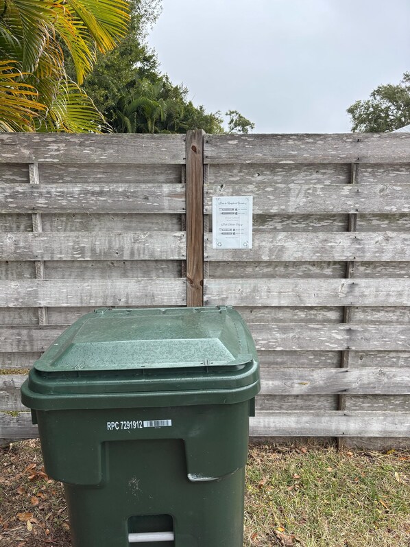 Un safataixa verd amb etiqueta de reciclatge i un codi de barres, col·locat davant d'una tanca de fusta amb un senyal de reciclatge.