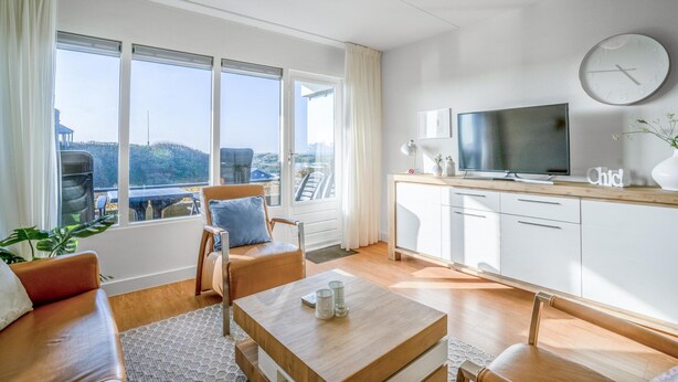 A modern living room with a sofa, coffee table, and TV. Large windows offer a view of the outdoors. - 4-person Apartment With Island-sea View (De Koog)