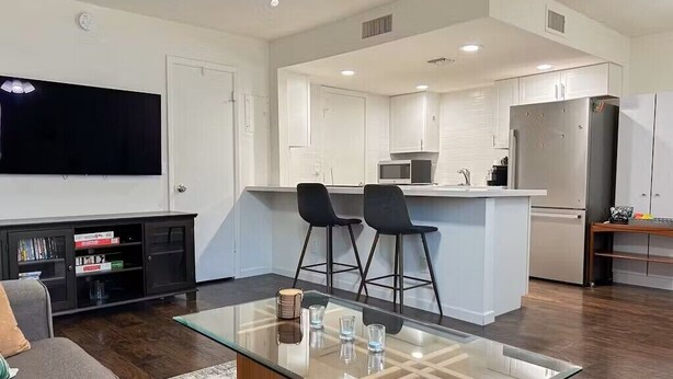 A modern kitchen with white cabinets, a stainless steel refrigerator, and a dining area with a glass table and two black bar stools.