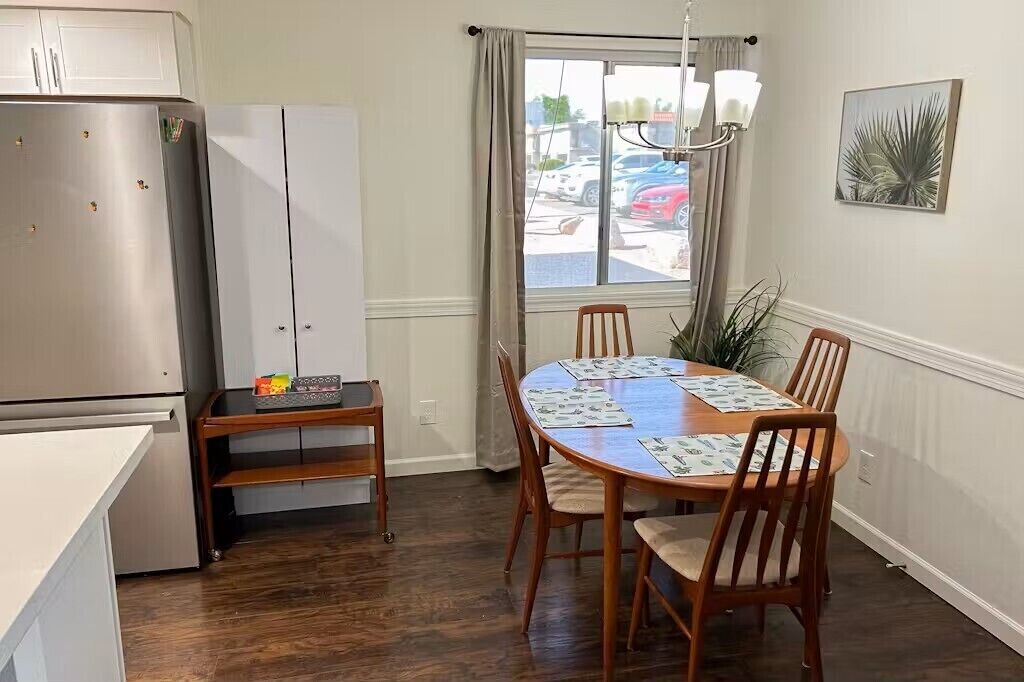 Una cocina compacta con refrigerador, una mesa de comedor con sillas y un ventanal con cortinas.