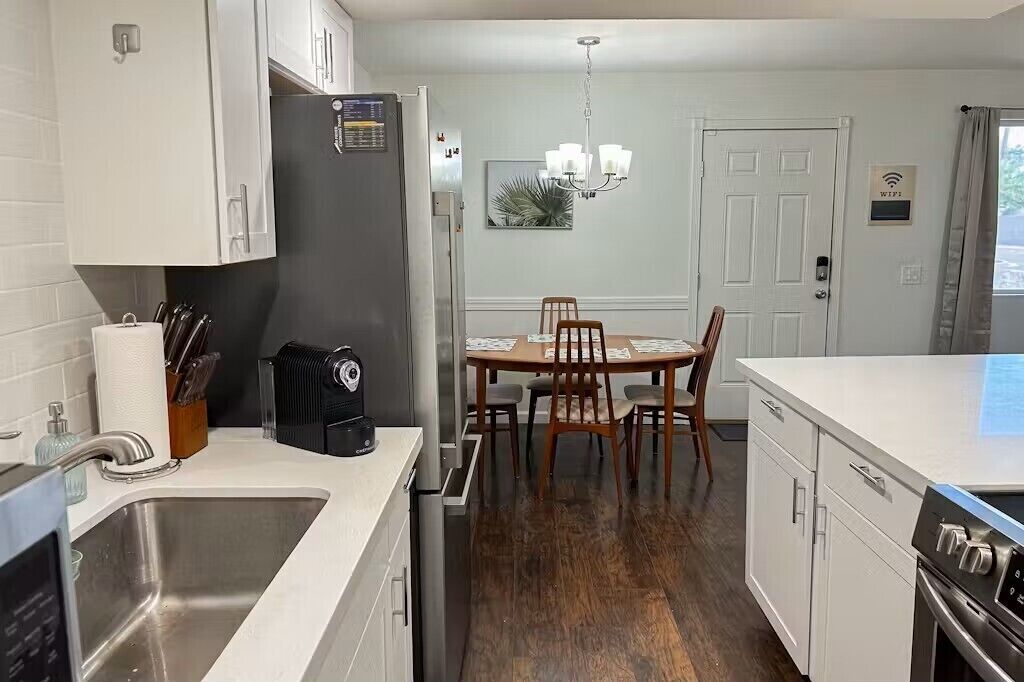 A modern kitchen with white cabinetry, a stainless steel sink, a black refrigerator, and a dining area with a wooden table and chairs.