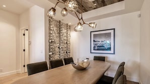 Uma sala de jantar com uma mesa de madeira, seis cadeiras, um candelabro, uma adega e um quadro na parede.