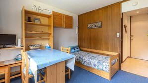 A compact room with a wooden desk, a small bed with patterned bedding, and a wall-mounted shelf with a television and decorative items. - Apartment in Plagne Centre by Ski Slopes (La Plagne-Tarentaise)