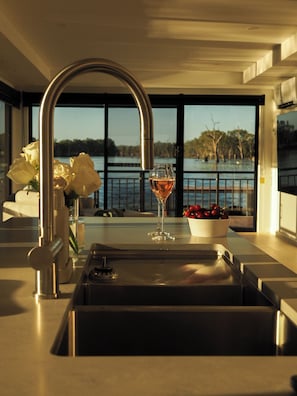 A modern kitchen with a stainless steel sink, a wine glass, and a bowl of fruit. - Odyssey - Luxury Houseboat (Renmark)